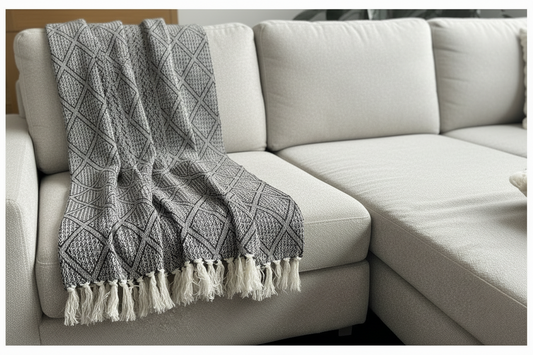 A woven throw blanket and matching square pillow with a black and white geometric pattern, styled on a light beige sectional sofa.