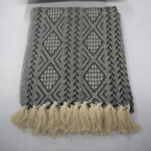 A woven throw blanket and matching pillow set with a black and beige geometric diamond pattern and cream fringe tassels, displayed on a light gray surface.