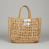 A natural woven straw tote bag with two top handles, laid flat on a dark gray tiled floor. A small white paper tag is attached to the front.