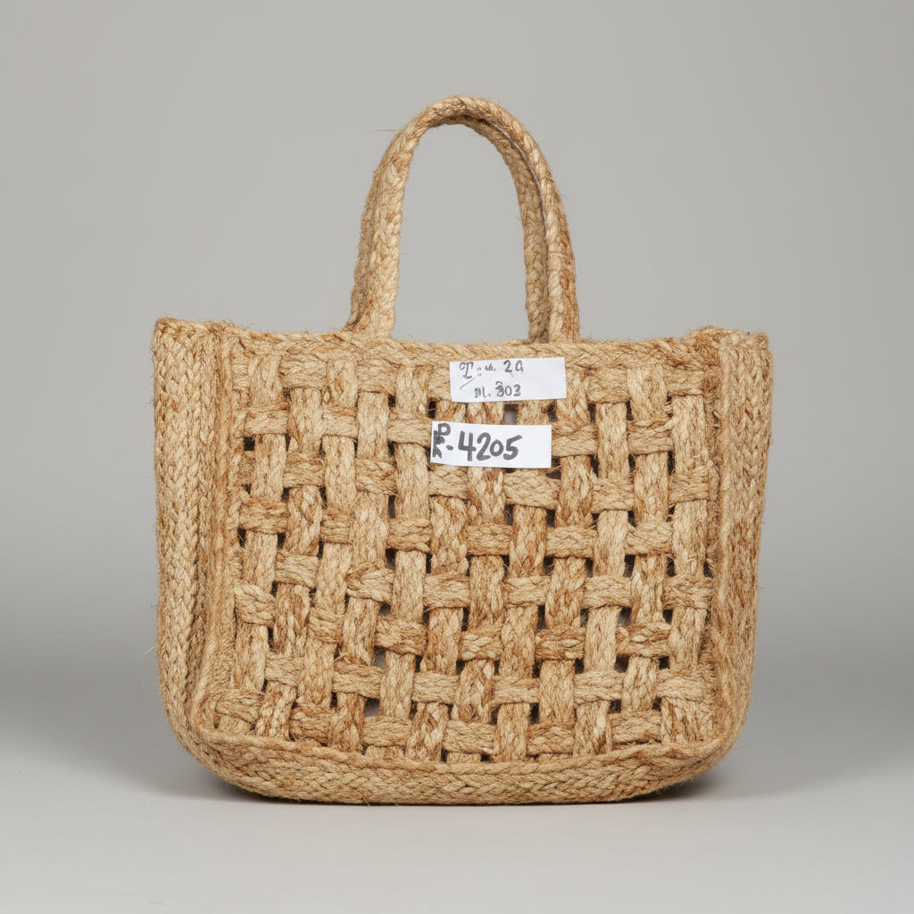 A natural woven straw tote bag with two top handles, laid flat on a dark gray tiled floor. A small white paper tag is attached to the front.